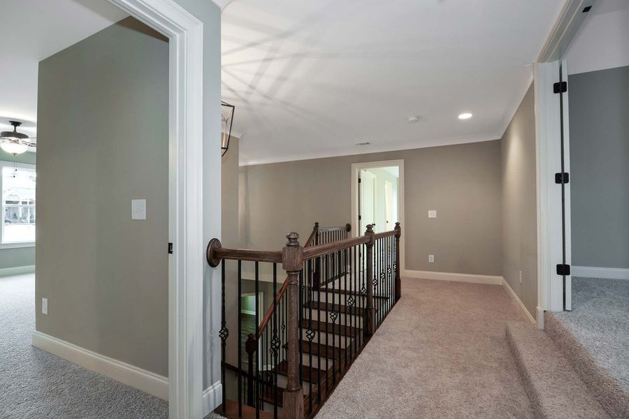 Hallway with stairs, light gray walls, beige carpet, and a dark wooden railing.