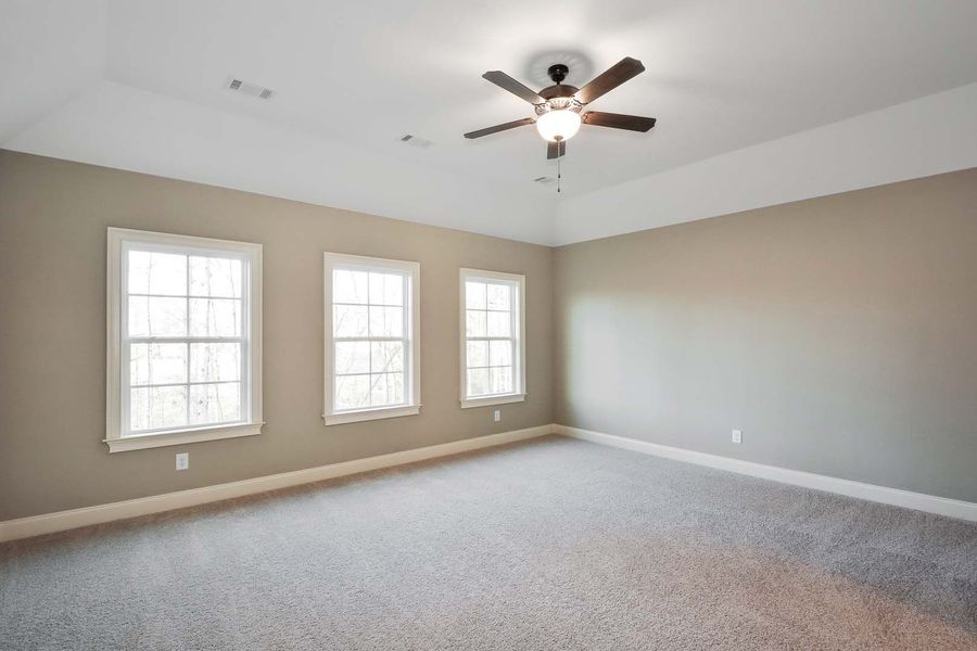 Empty bedroom with beige walls, three windows, gray carpet, and ceiling fan.