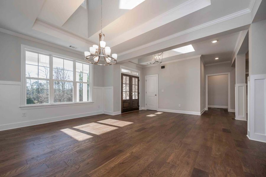 Empty, bright dining room with dark wood floors, white trim, large windows, and chandeliers.