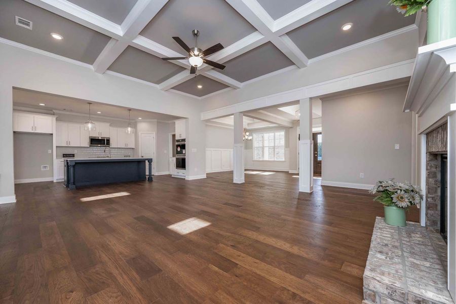 Spacious living area with dark wood floors, coffered ceiling, kitchen in the background, and a fireplace.
