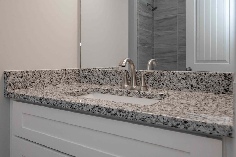 White vanity with a granite countertop, sink, and faucet. Mirror and shower in background.
