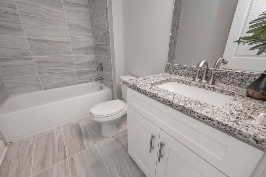 Bathroom with gray-tiled walls and floor, white vanity with granite countertop, white tub, and toilet.