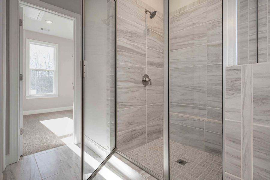 Bathroom with a glass shower, gray tiles, and a glimpse into a bedroom with sunlight.