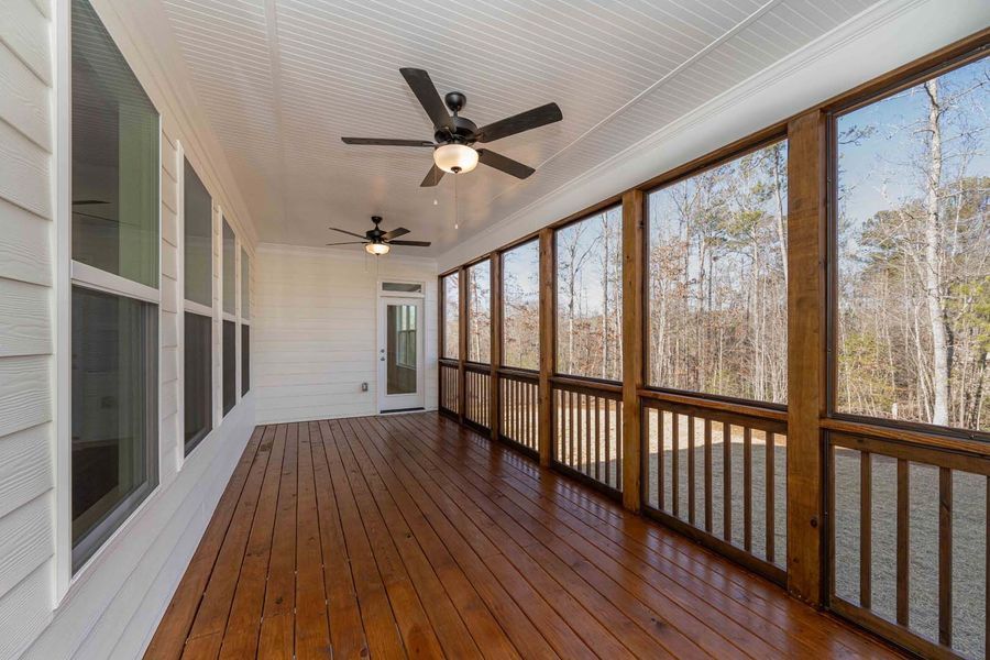 A screened porch with wooden floor, railing, ceiling fans, and large windows overlooking a wooded area.