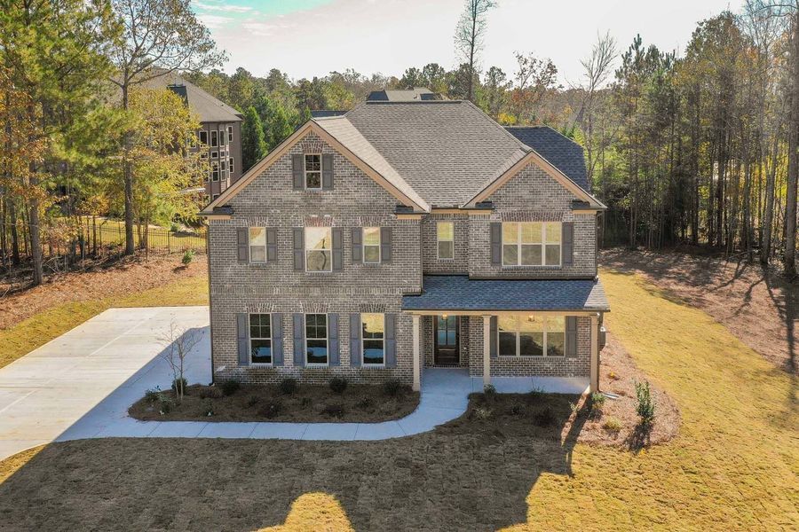 Two-story brick house with gray roof, front porch, and concrete driveway, surrounded by trees.