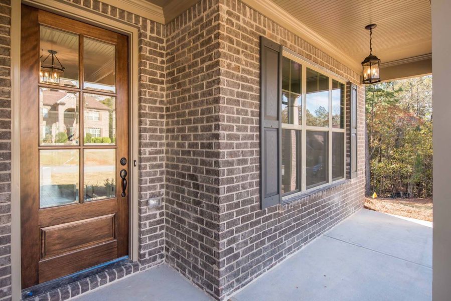 Brick-walled house entrance with a wooden door, windows, dark shutters, and a covered porch.