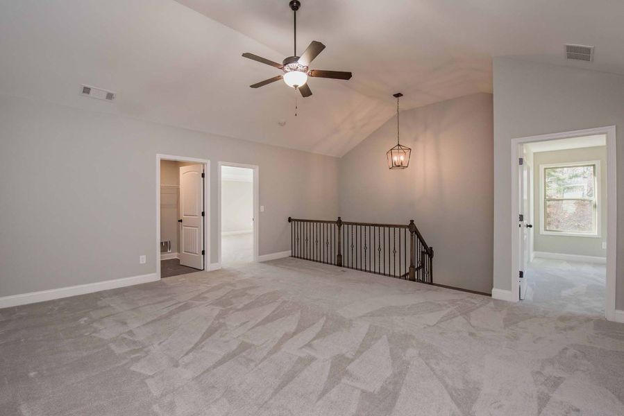 Spacious room with gray carpet, walls, and a sloped ceiling. A dark staircase, ceiling fan, and chandelier are visible.