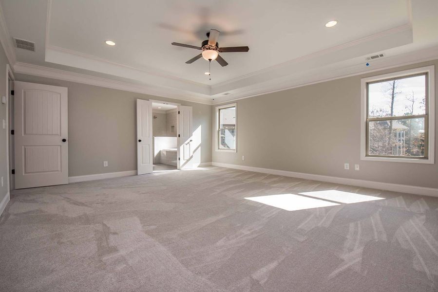 Empty bedroom with beige walls, carpet, and a ceiling fan. Two windows allow sunlight in.