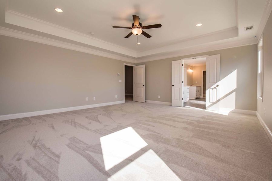 Empty bedroom with beige walls, carpet, and ceiling fan, bright sunlight through a window.