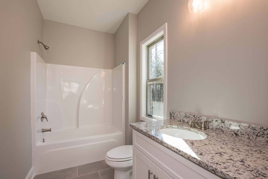 Bathroom with white tub, toilet, and vanity, gray walls, window, and granite countertop.