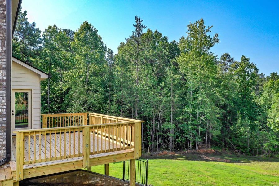 Wooden deck attached to a house, overlooking a grassy yard and a line of trees under a blue sky.
