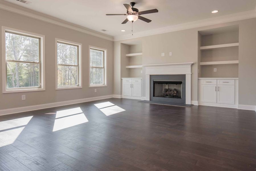 Empty living room with dark wood floors, fireplace, built-in shelves, and three windows.
