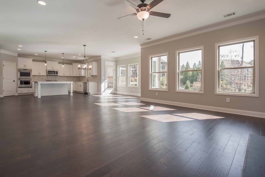 Empty, open-concept living space with dark wood floors, light walls, and kitchen in the background.