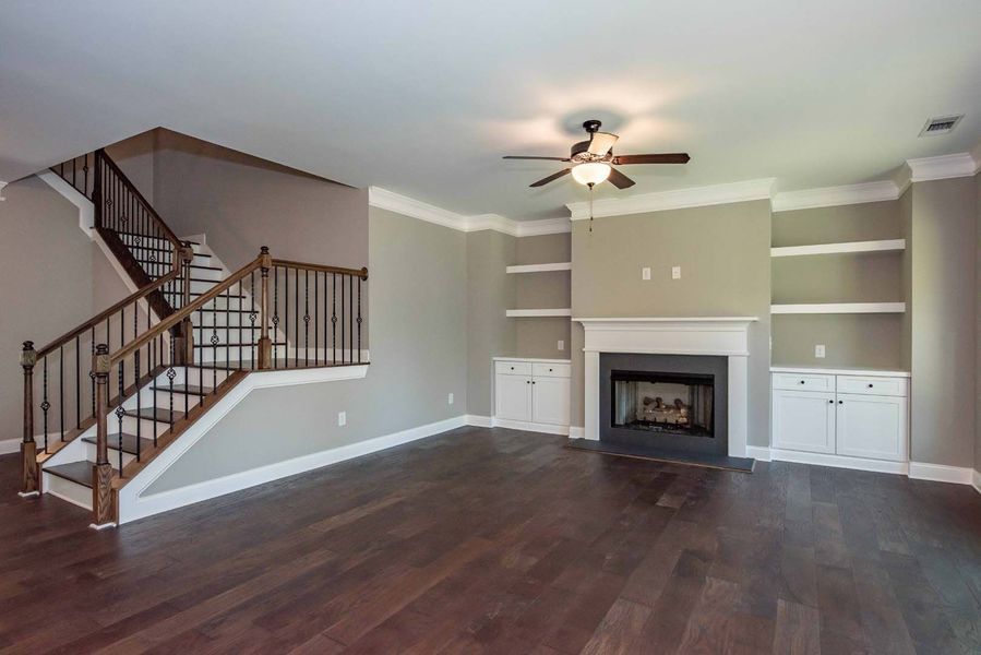 Living room with a fireplace, built-in shelves, and a staircase. Dark wood floors, neutral walls, and a ceiling fan.
