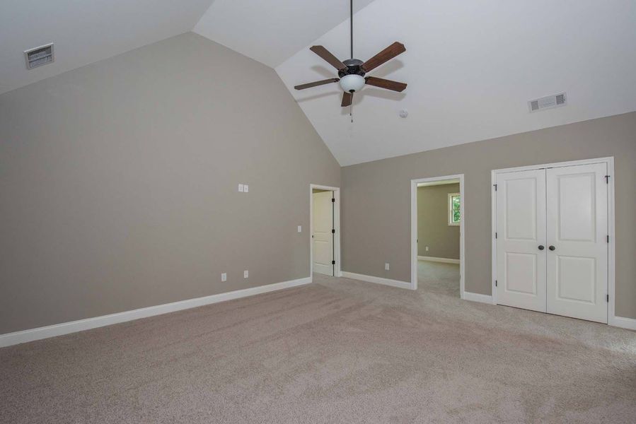 Empty bedroom with vaulted ceiling, beige carpet, gray walls, and white doors. Ceiling fan.
