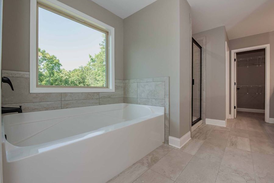 Bathroom with a white bathtub, window, and shower, gray walls, and light gray tile flooring.