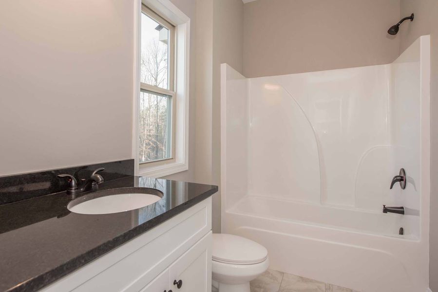 Bathroom with white tub/shower, toilet, vanity with black countertop, and a window.