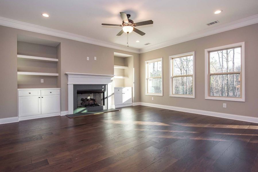 Empty living room with fireplace, built-in shelves, and hardwood floor. Three windows.