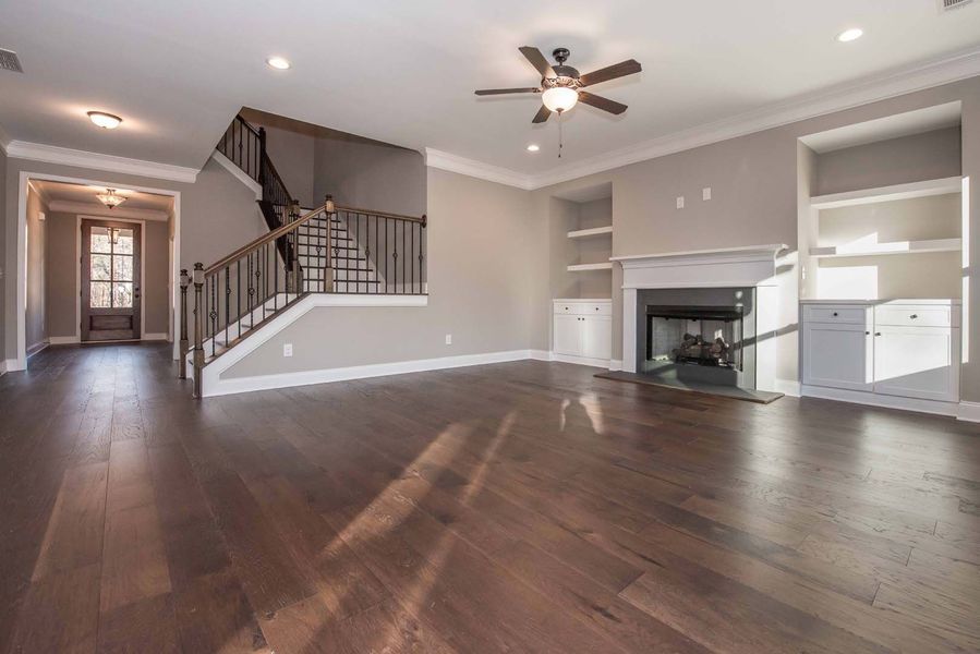 Spacious living room with dark wood floors, fireplace, and staircase, lit by natural light.