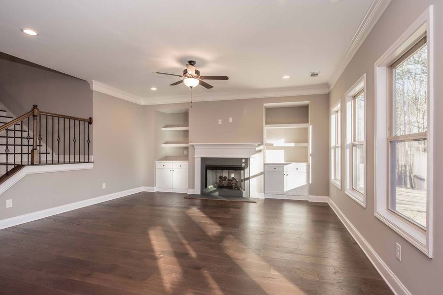 Empty living room with fireplace, built-in shelves, windows, dark wood floor, and a ceiling fan.