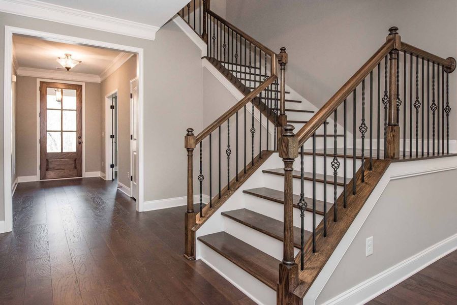 Interior view of a home's entry hall with a staircase. Wooden floors, beige walls, and dark wood and iron railing.