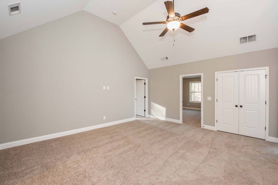 Empty bedroom with vaulted ceiling, tan carpet, gray walls, white trim and doors, and a ceiling fan.