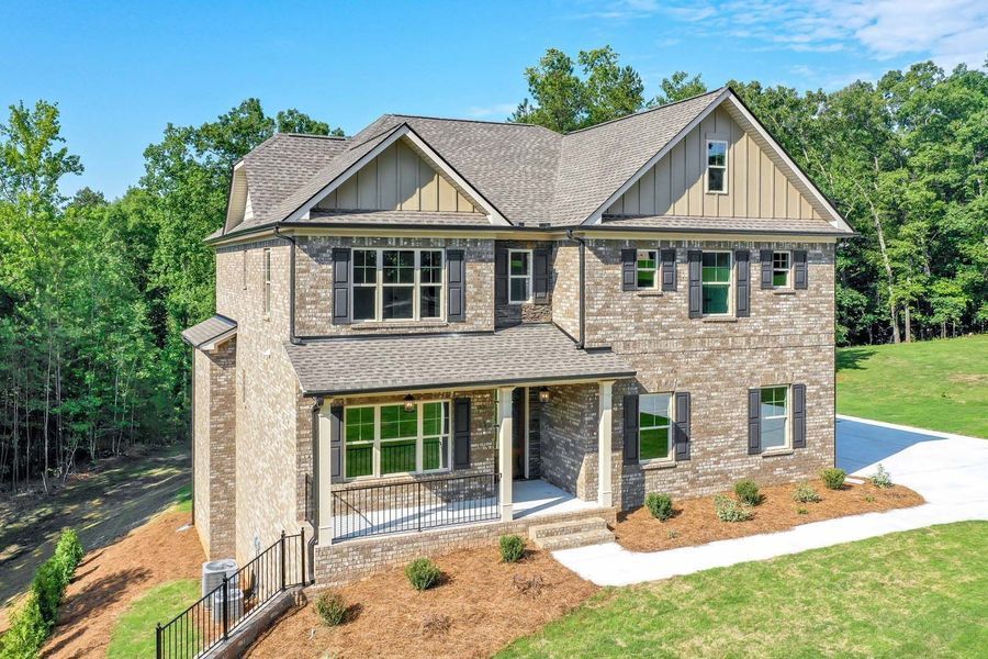 Two-story brick house with brown roof and shutters, surrounded by trees and grass on a sunny day.