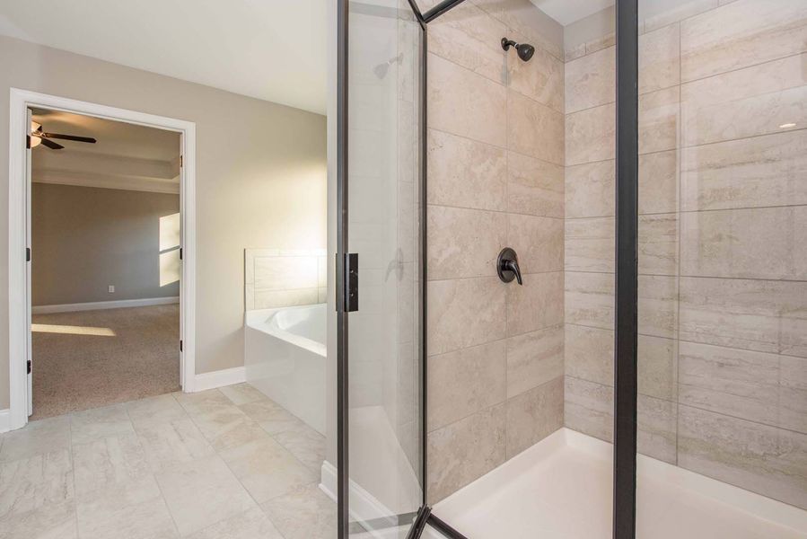 Bathroom with glass shower, tan tiles, and view into bedroom with neutral tones.