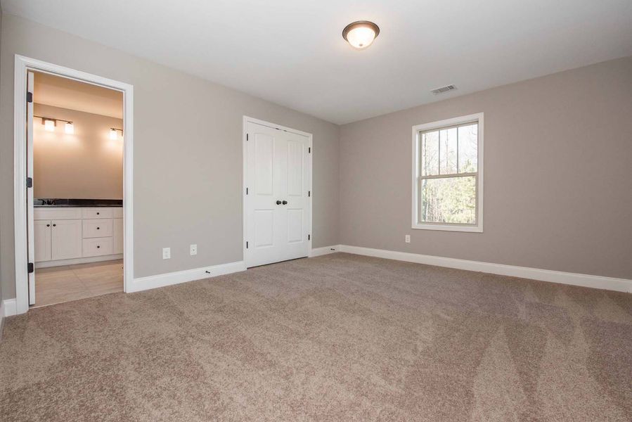 Empty bedroom with gray walls, beige carpet, white trim, and a door to a bathroom.