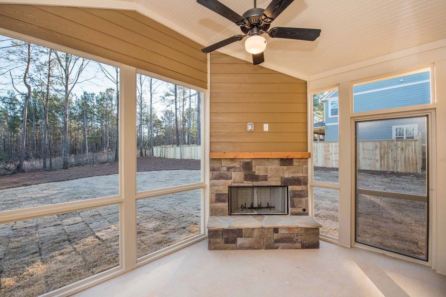 Sunroom with stone fireplace, windows overlooking a yard and trees. Brown, beige, and white color scheme.
