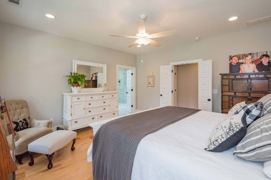Bedroom with a bed, dresser, chair, and bathroom door.  White walls, light wood floors, and a ceiling fan.