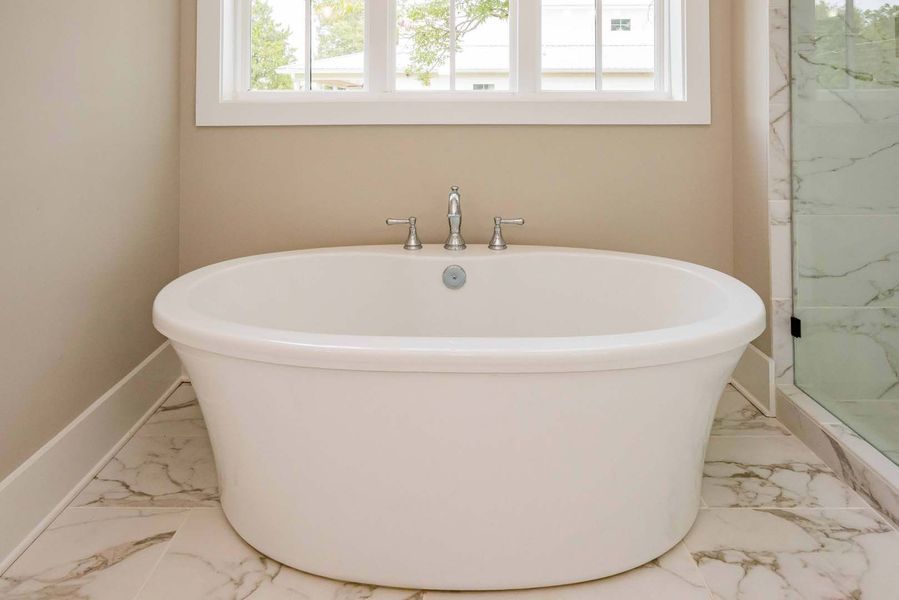 White oval bathtub beneath a window in a bathroom with marble tile and beige walls.