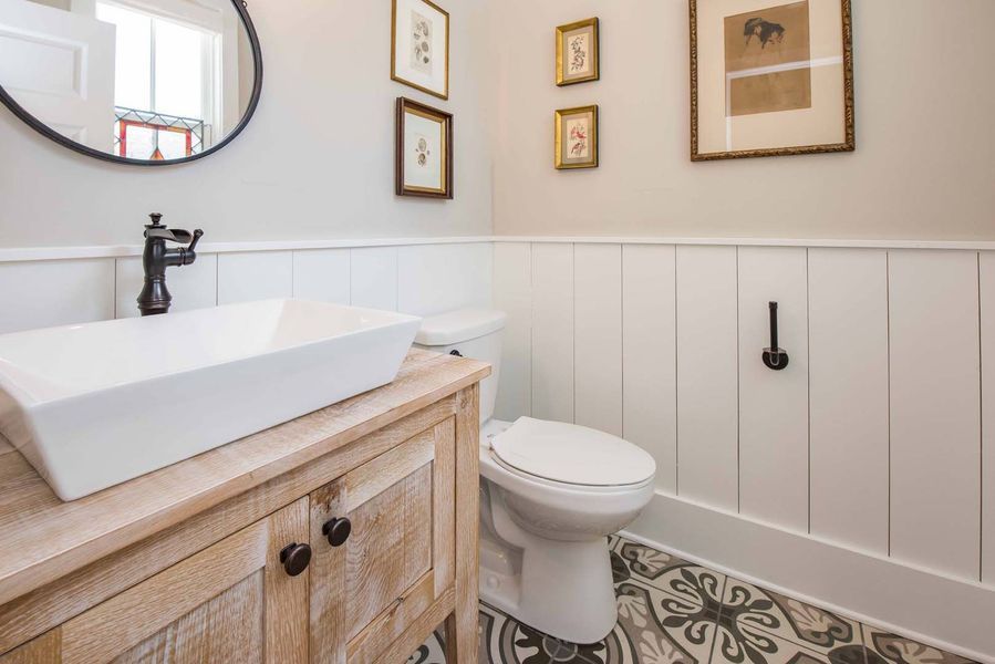 Powder room with a white vessel sink on a light wood vanity, white wainscoting, and patterned floor tiles.