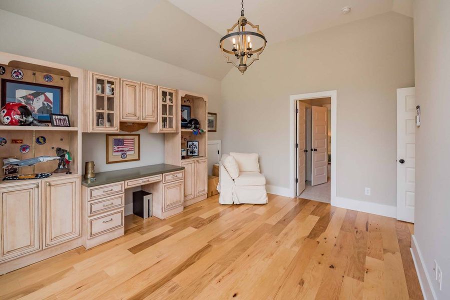 Home office with light wood floors, built-in cabinets, desk, armchair, and open doorway.