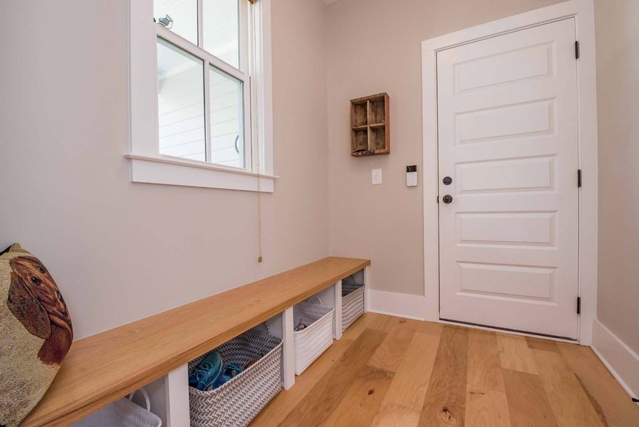 Mudroom with wooden bench, baskets, white door, and window. Neutral walls and wood floor.