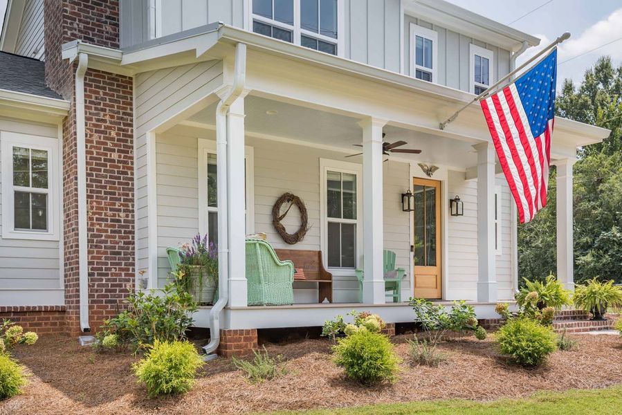 Farmhouse with porch, American flag, and potted plants. Light gray siding and brick chimney.
