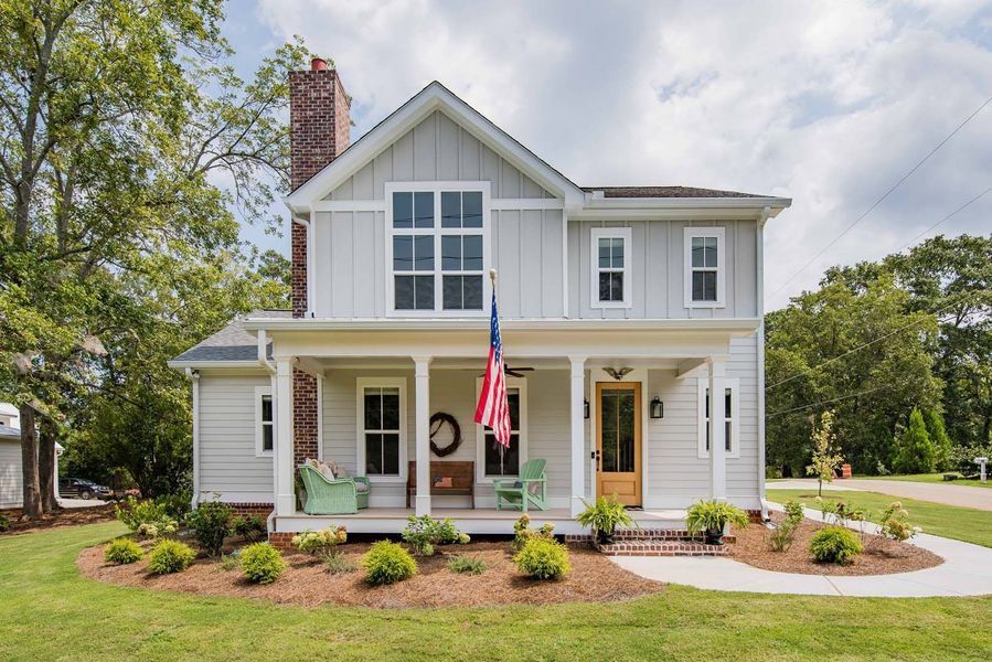 Two-story light grey house with porch, American flag, and brick chimney, on a grassy lawn.