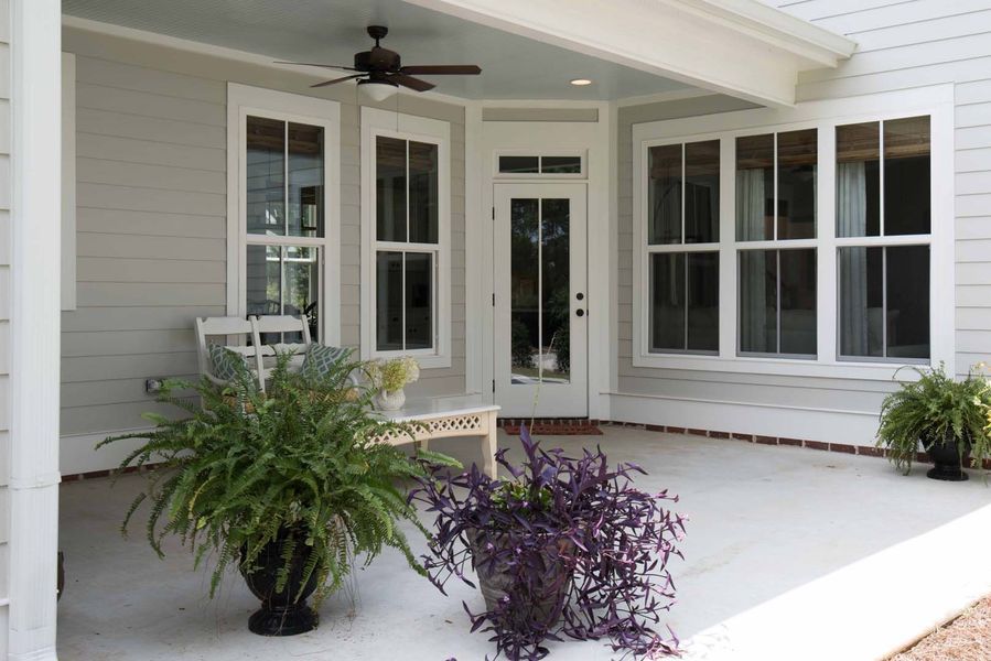 Covered porch with white-framed windows, a door, ferns, and a bench against a light gray building.