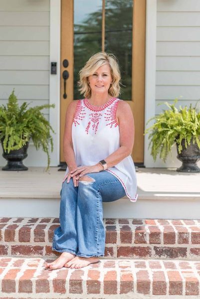Blonde woman in jeans sits on brick steps, wearing a white and pink top, in front of a house.