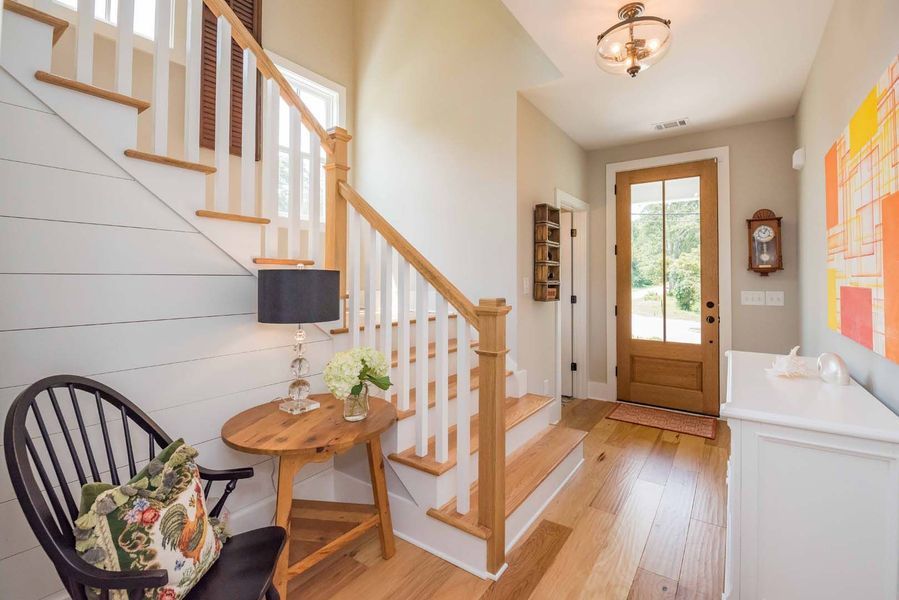 A bright entryway with wooden stairs, a small table, a chair, and a front door.