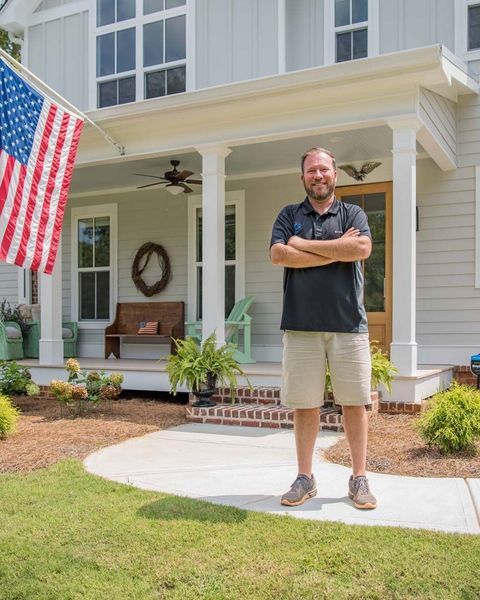 Man standing in front of a light gray house with an American flag. He's smiling, arms crossed.