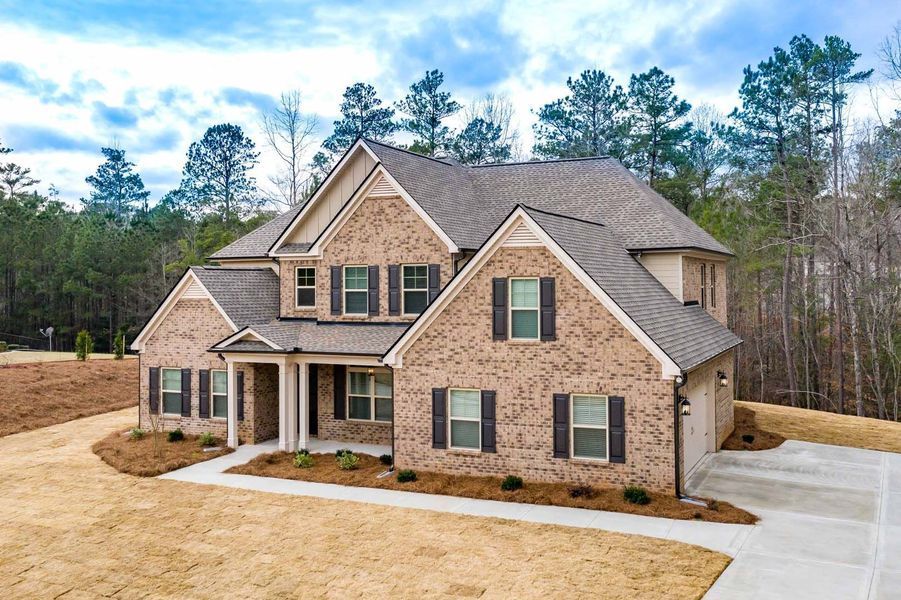Two-story brick house with brown roof and shutters, front porch, driveway, and trees in the background.