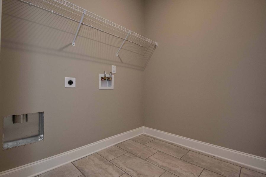 Empty laundry room with beige walls, tiled floor, and wire shelf above washer/dryer hookups.