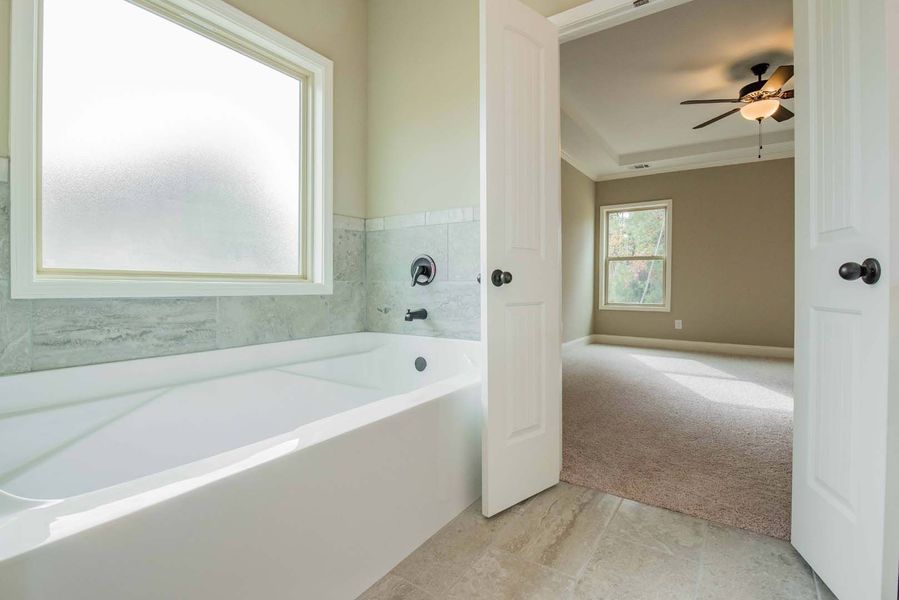 Bathroom with white bathtub, window, and doorway to a bedroom with carpet and ceiling fan.