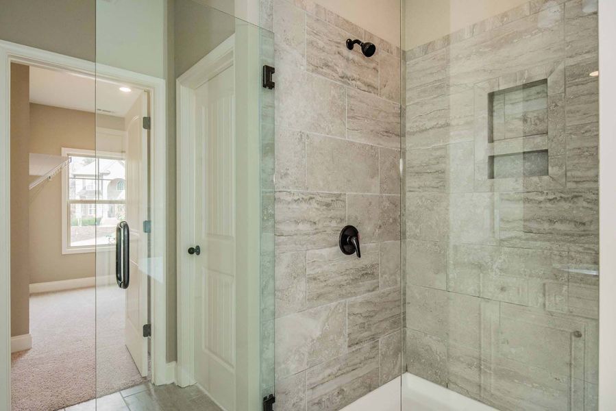 Bathroom with glass shower and door, light grey tile walls, black fixtures, and a doorway to a bedroom.