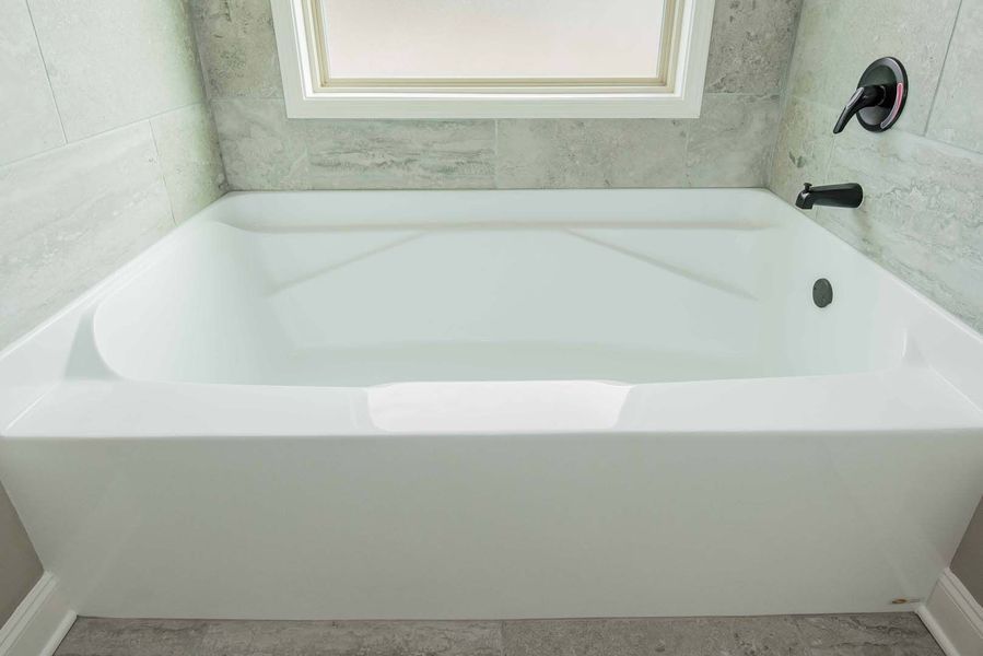 White built-in bathtub with black faucet, beneath a small window. Gray tiled walls and floor.