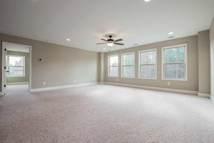 Empty, spacious room with beige walls and carpet. Large windows let in light; a ceiling fan hangs in the center.