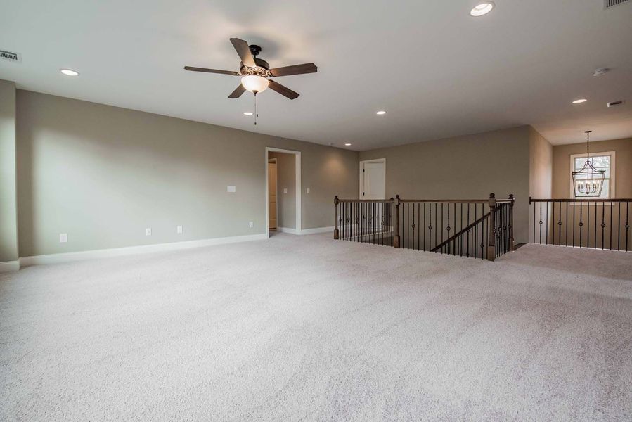 Large room with tan walls, white carpet, and a dark ceiling fan. A railing and stairs are visible.