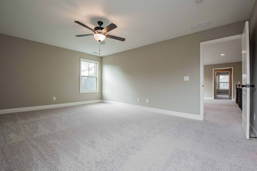 Spacious, empty bedroom with gray walls, light gray carpet, and a ceiling fan. Doorway leads to a hallway.