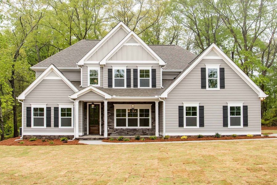 Two-story light gray house with black shutters, stone accents, and a brown door in a yard with trees.